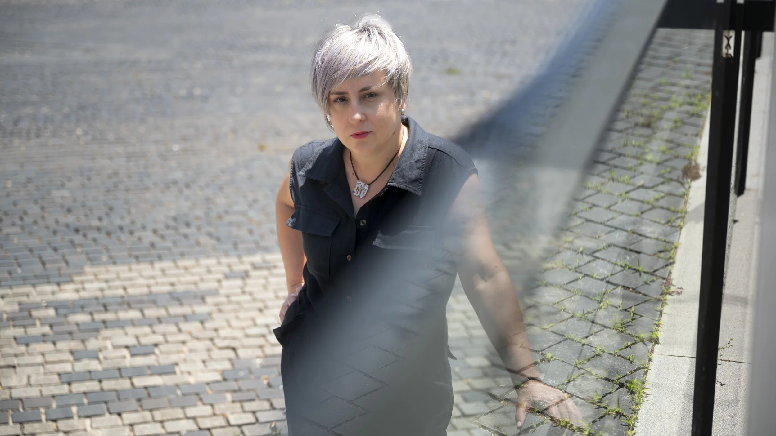 Mujer con cabello corto y plateado, con camisa negra, mostrando una expresión seria, sentada en una banca en la calle.