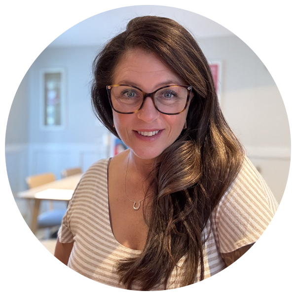 Sundy, with glasses on, smiling at the camera in a room with a white wall and a soft blur background.