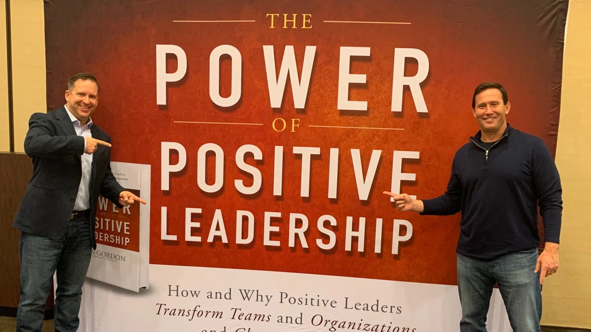 Two men smiling and pointing at a large banner displaying "The Power of Positive Leadership."