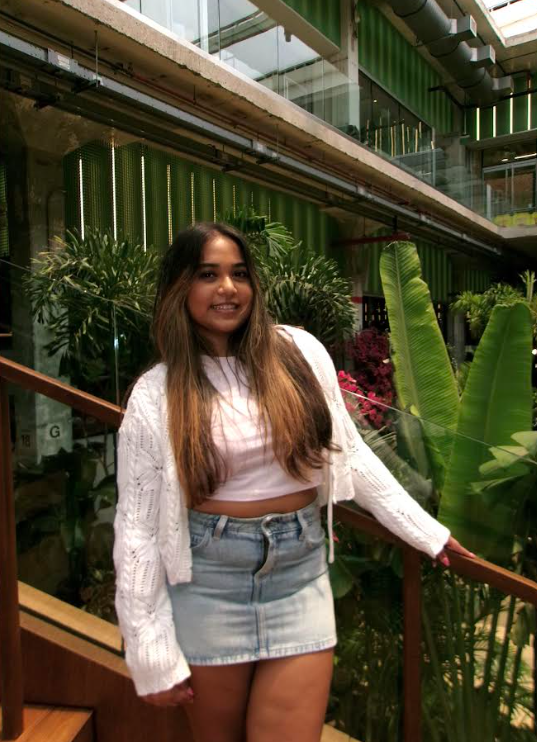 Young woman with long brown hair smiling, wearing a white crop top, white cardigan, and light denim skirt, standing indoors among lush green plants and glass railings.