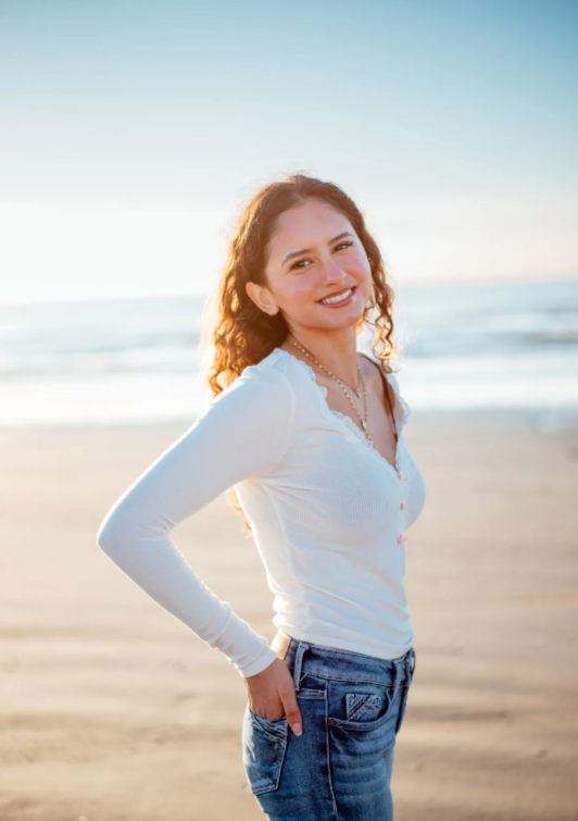 A woman standing on a beach during sunset, smiling at the camera with her hand on her hip.