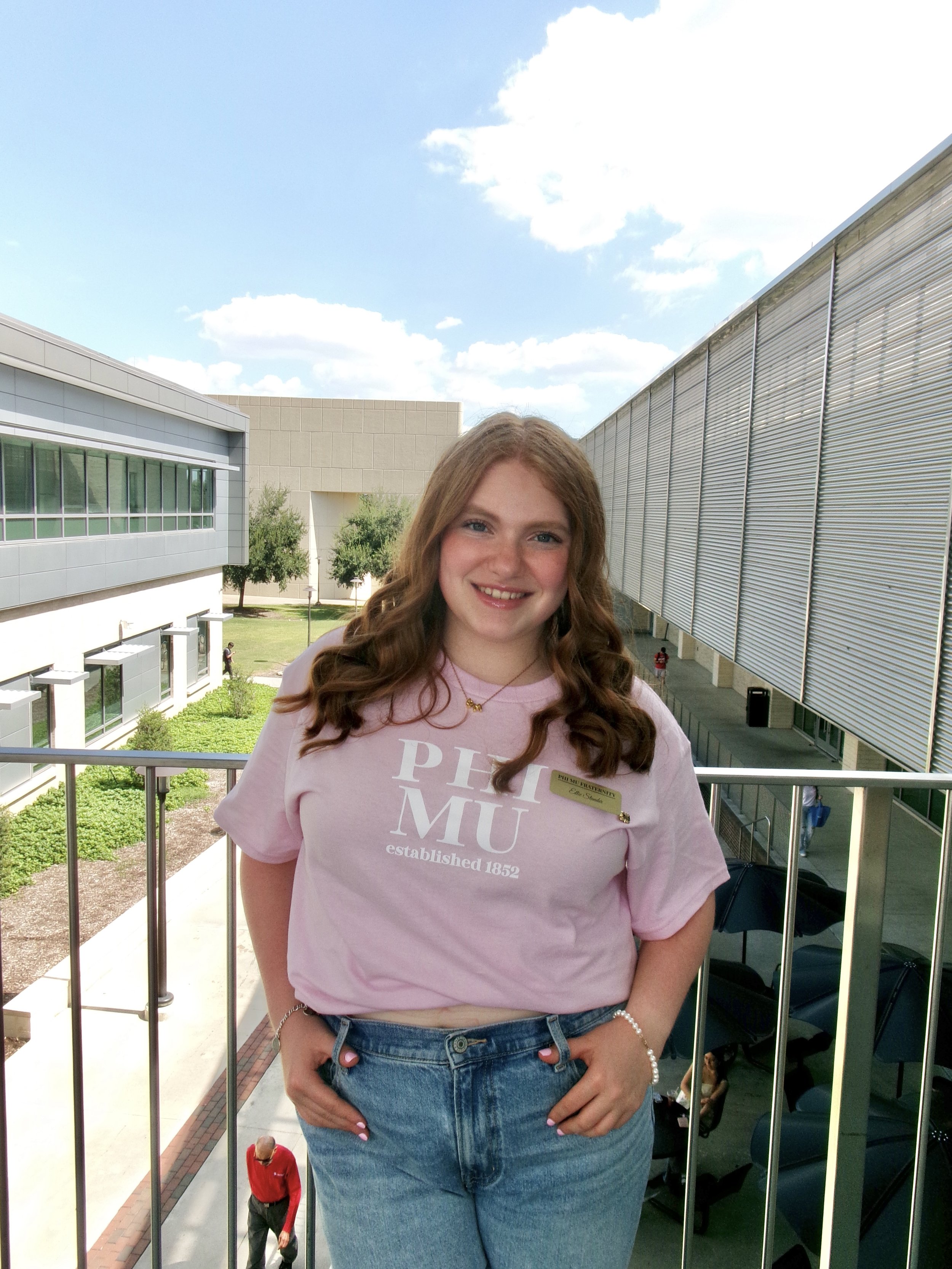 A young woman with long red hair smiling on a university campus balcony, wearing a pink Pitt MUSE T-shirt, jeans, with a name tag and a pearl bracelet.