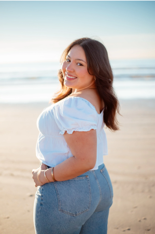 Woman smiling at the beach during sunset, wearing a white top and blue jeans.