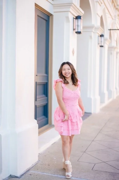 A woman in a pink dress standing on a sidewalk in front of a light-colored building with blue doors and black wall lamps.