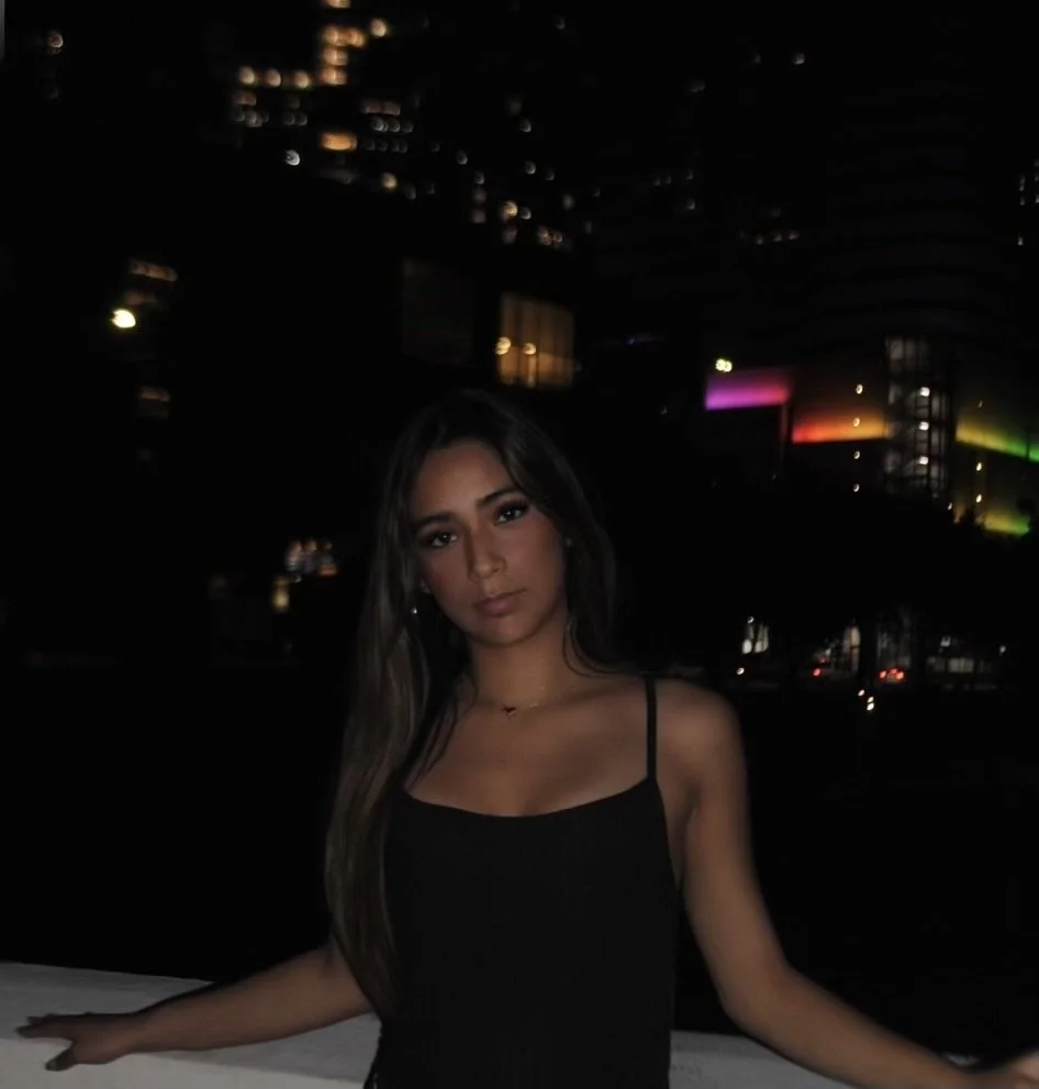 A young woman with long dark hair taking a nighttime photo outdoors with city lights and buildings in the background.