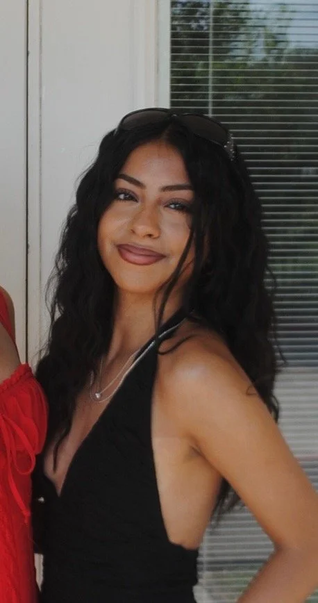 A woman with long, curly black hair, wearing sunglasses on her head, a black sleeveless top, and a necklace, smiling in front of a window with blinds.
