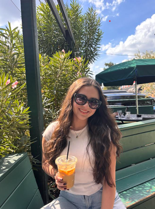 A young woman with long wavy hair wearing sunglasses and a white t-shirt, holding a cold drink with a straw, sitting outdoors at a cafe with green lupines and a dark green umbrella in the background.
