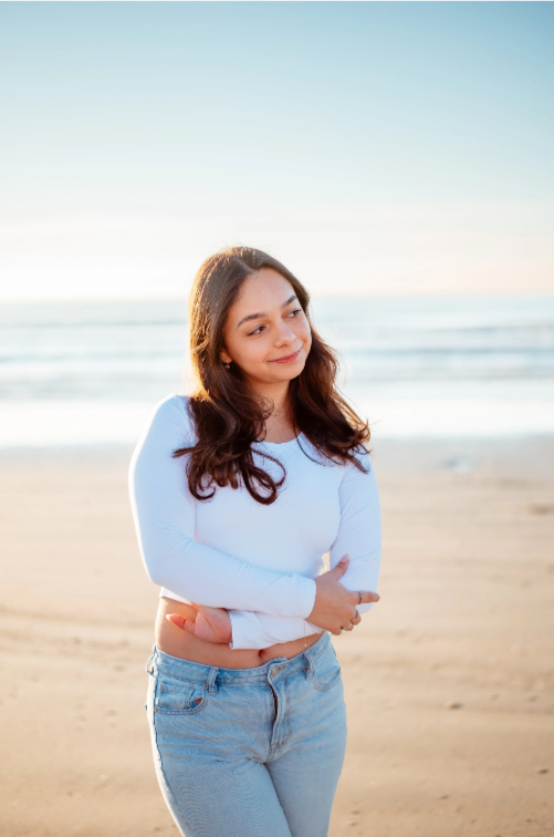 Young woman with long hair wearing a white long-sleeve shirt and jeans standing on a beach during sunset.