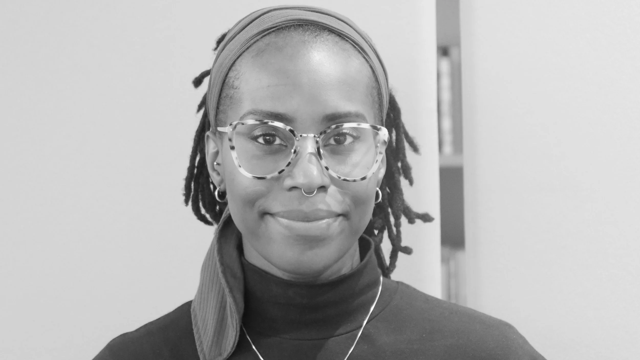 Black-and-white photo of a Black woman with spotted glasses, a head scarf over  black locs and a closed-mouth smile.
