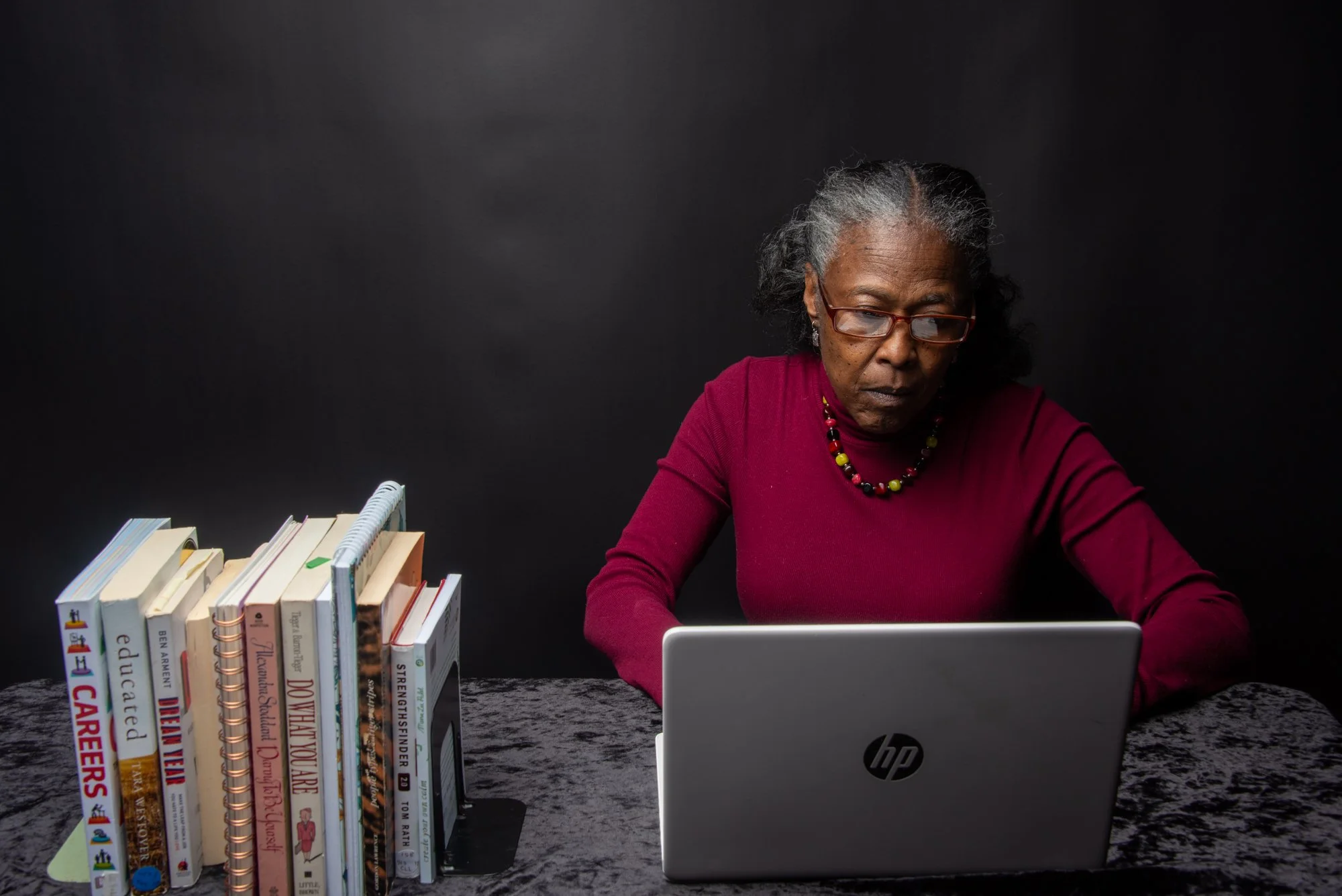 A woman in a red sweater working on an HP laptop at a desk. A collection of books is visible on the left side of the desk. The background is black.