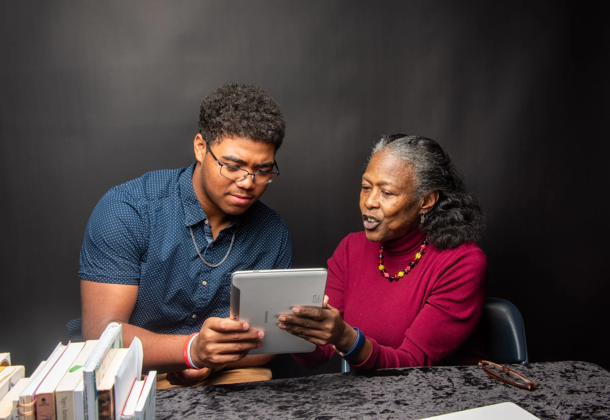 Older woman and young man looking at a tablet at a table with books.