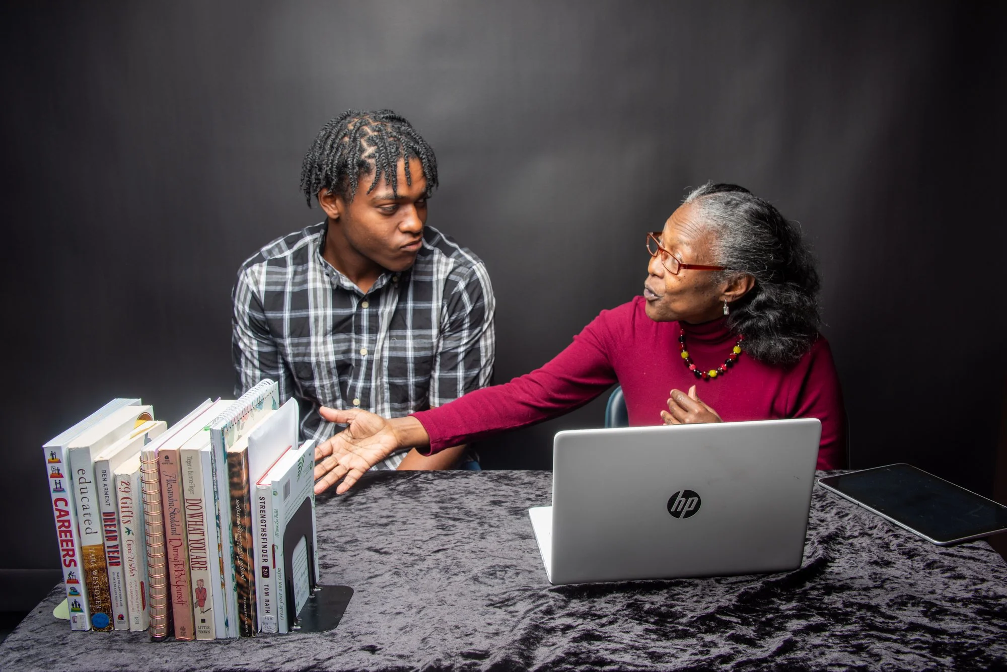 Two people sitting at a table with books and a laptop, having a discussion in front of a black background.