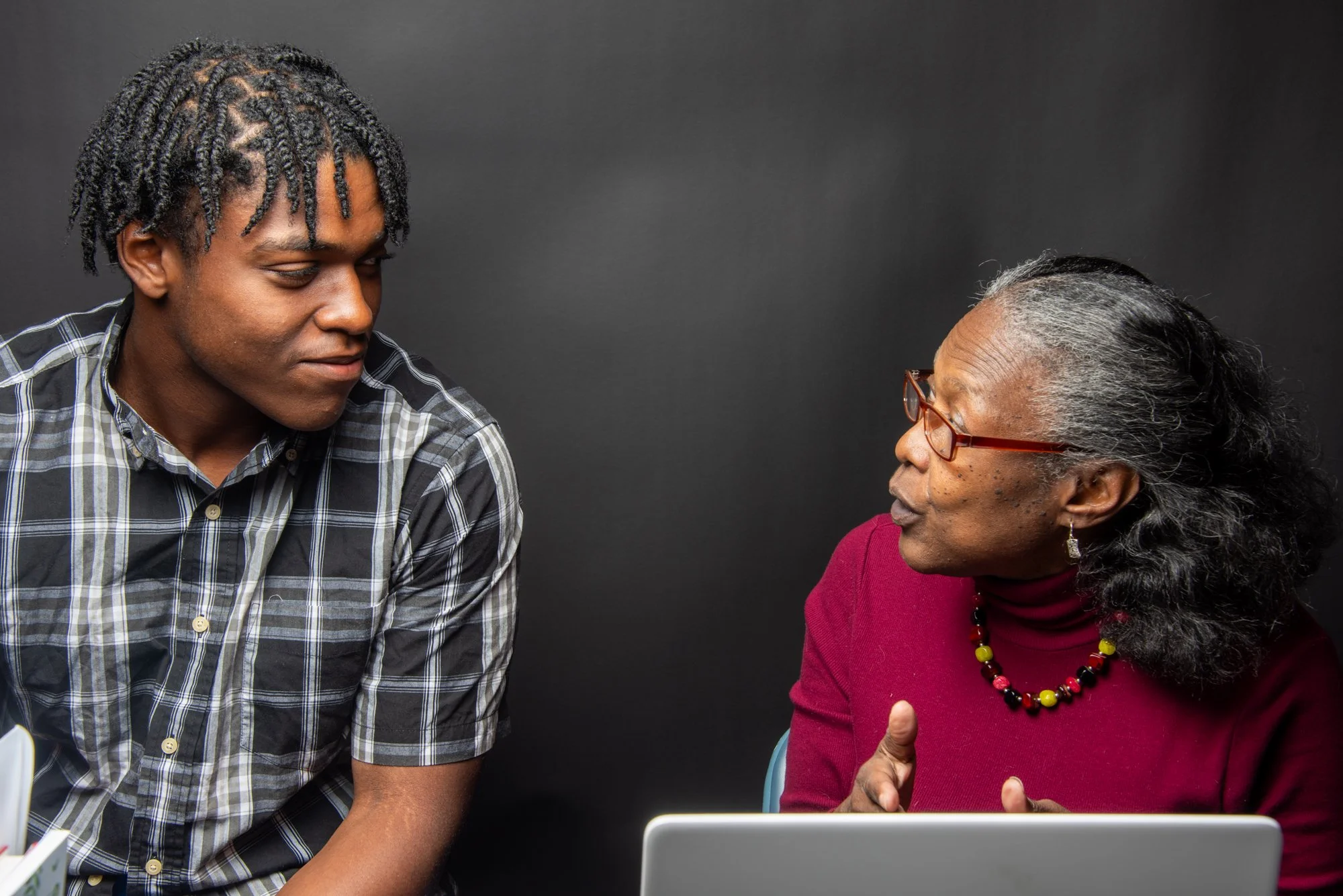 An older woman and a younger man sitting and talking, with the woman gesturing. She is wearing a red turtleneck and glasses, while the man is in a plaid shirt. There's a laptop in front of them.