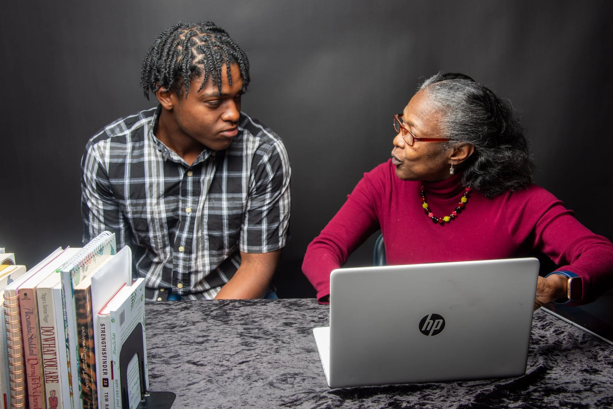 A young man and an older woman sitting at a table with books and a laptop, engaged in conversation. The man wears a plaid shirt, and the woman wears a burgundy sweater and glasses.