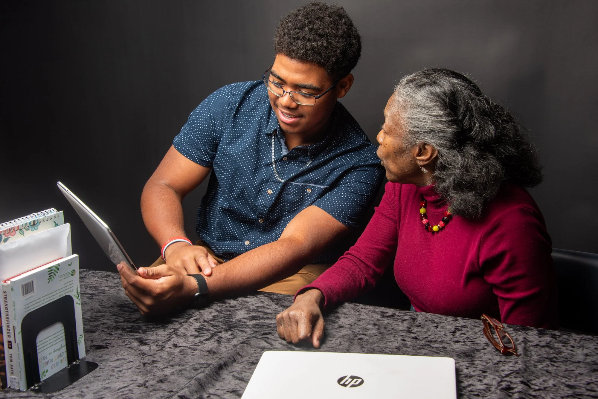 A young man showing a senior woman something on a tablet. They are sitting at a table with a laptop, a notebook, and a book to the side. The background is dark, and they appear engaged in conversation.
