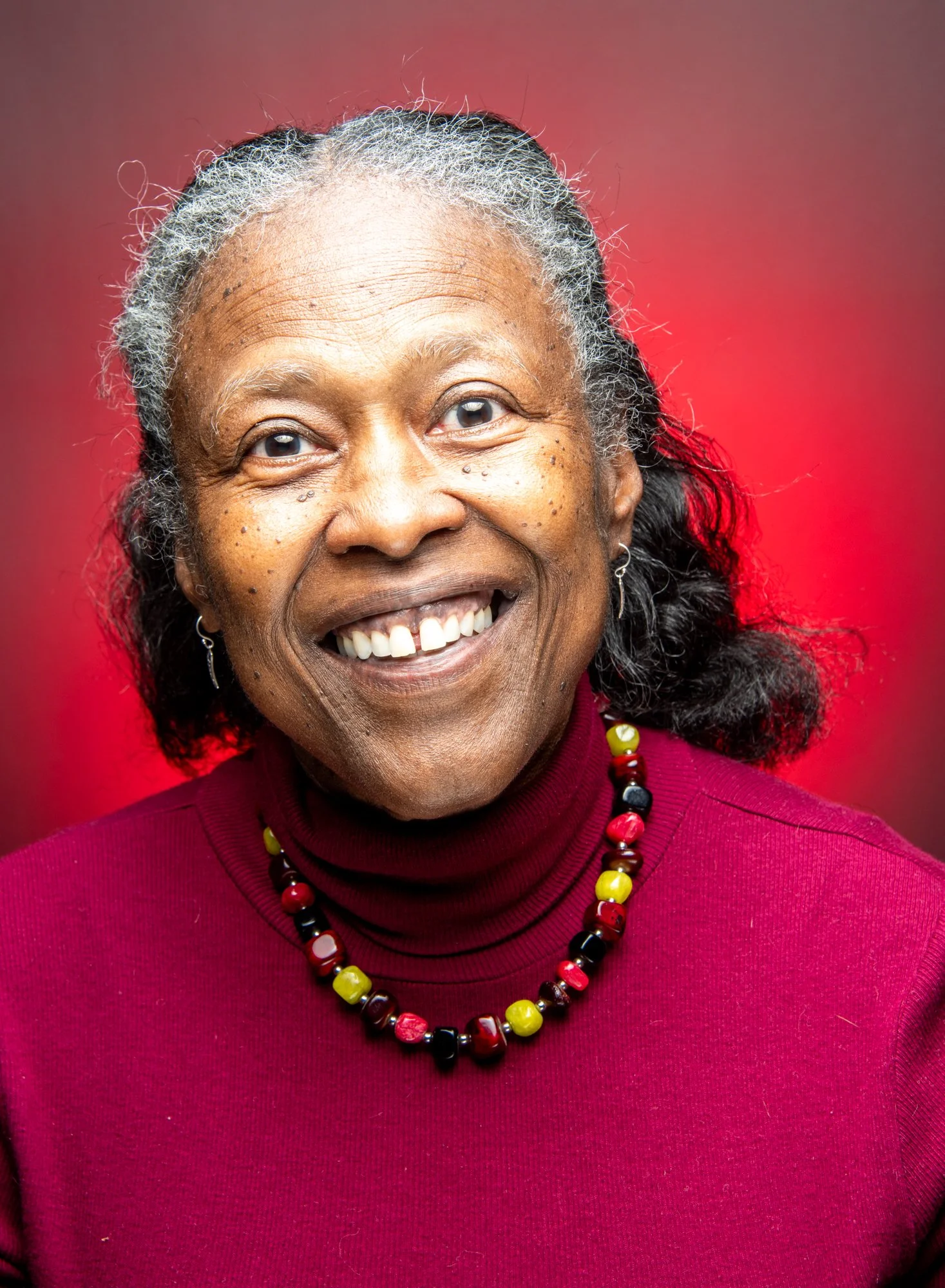 Elderly woman smiling with a red background, wearing a maroon sweater and a colorful bead necklace.