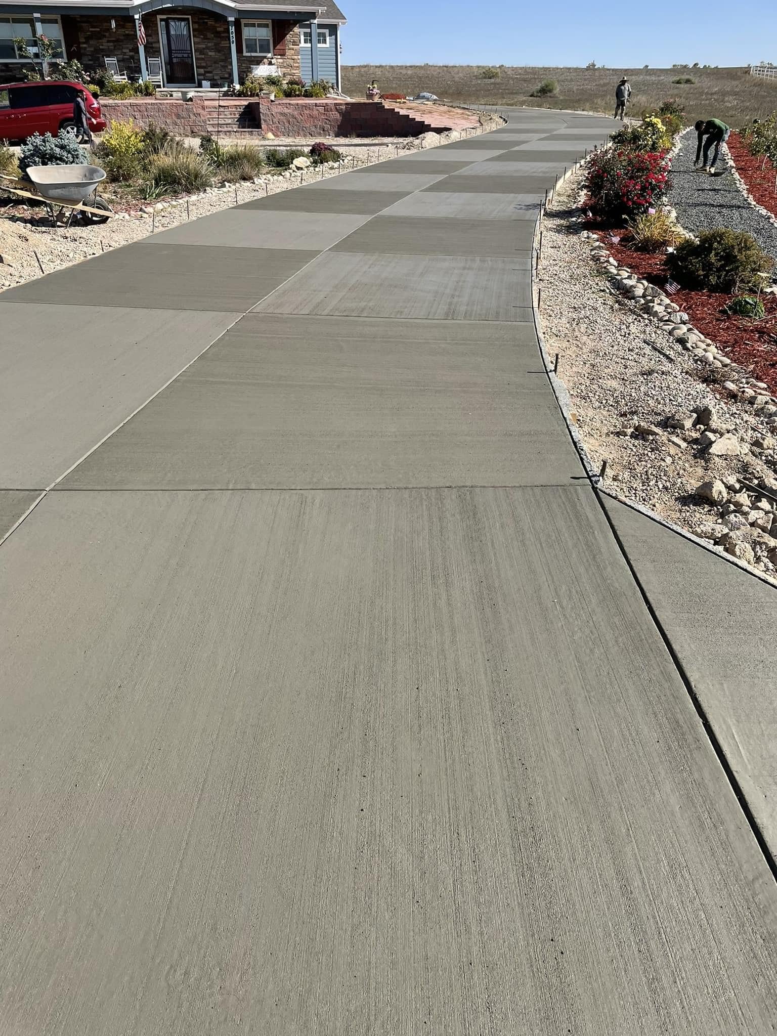 Newly laid concrete driveway leading to a house with a landscaped garden, workers in the background, and a wheelbarrow on the side.