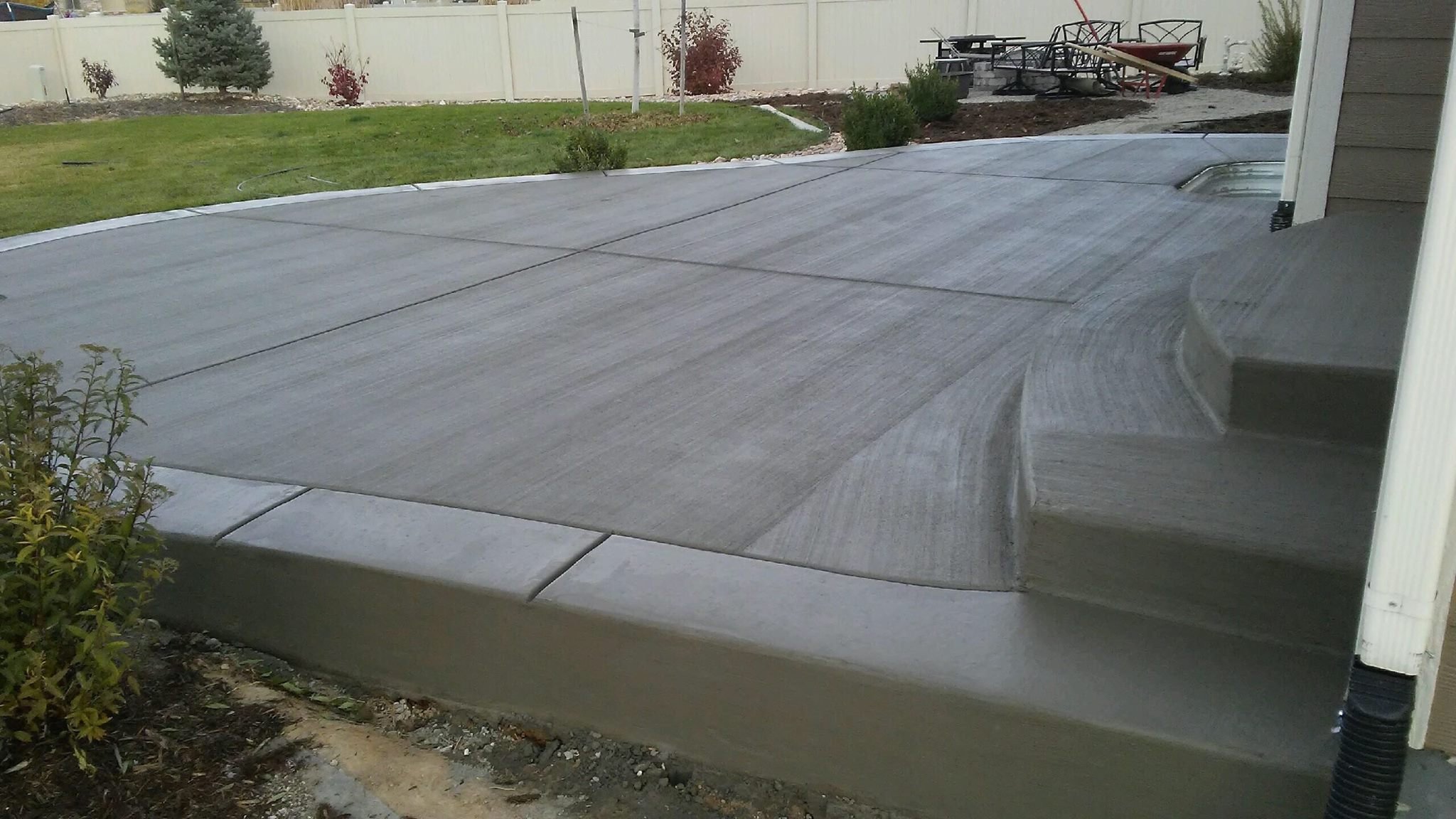 Freshly poured concrete patio with steps in a backyard, surrounded by grass and plants.