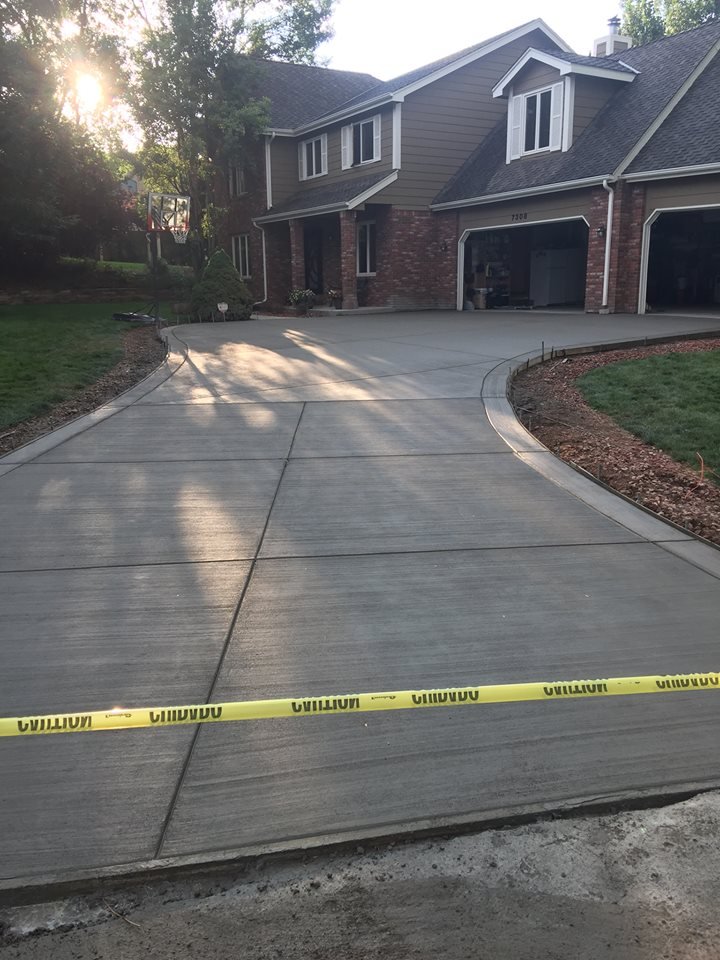 Newly poured concrete driveway with caution tape, leading up to a large suburban house with brick and siding exterior, featuring a two-car garage and surrounded by green lawn and trees.