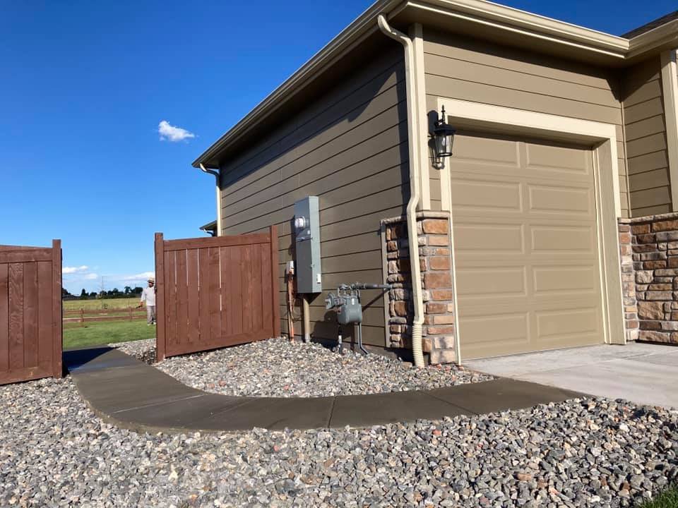 Exterior of a beige house with a brown wooden fence, a closed garage door, stone siding, a utility meter, and a gravel path on a sunny day.