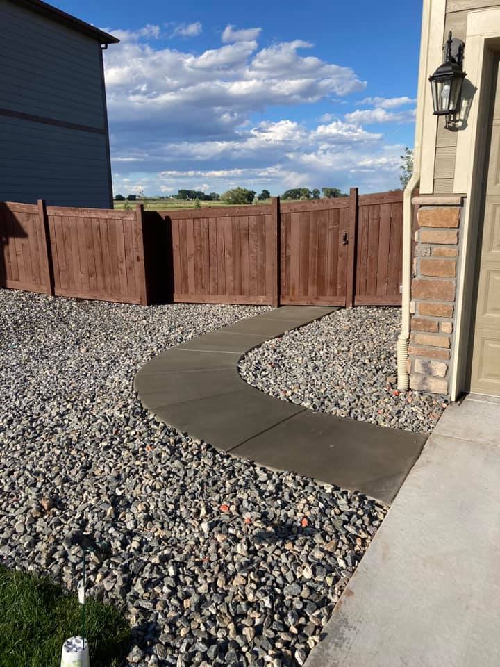 Curved concrete path through gravel yard, wooden fence, house wall in background, lantern-style outdoor light, blue sky.