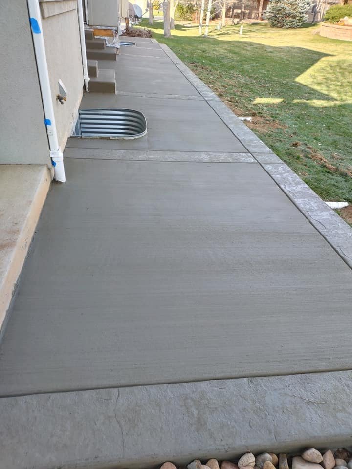 Freshly poured concrete patio alongside a house with a window well, yard, and downspout.