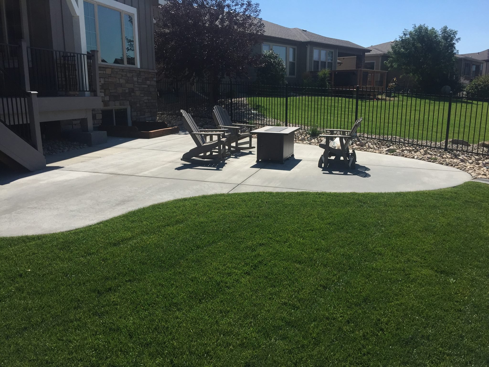 Backyard patio with Adirondack chairs and table, surrounded by grass and fenced garden area.