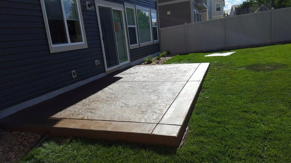 Backyard with a patio and grassy lawn next to a house with sliding doors and a fence.