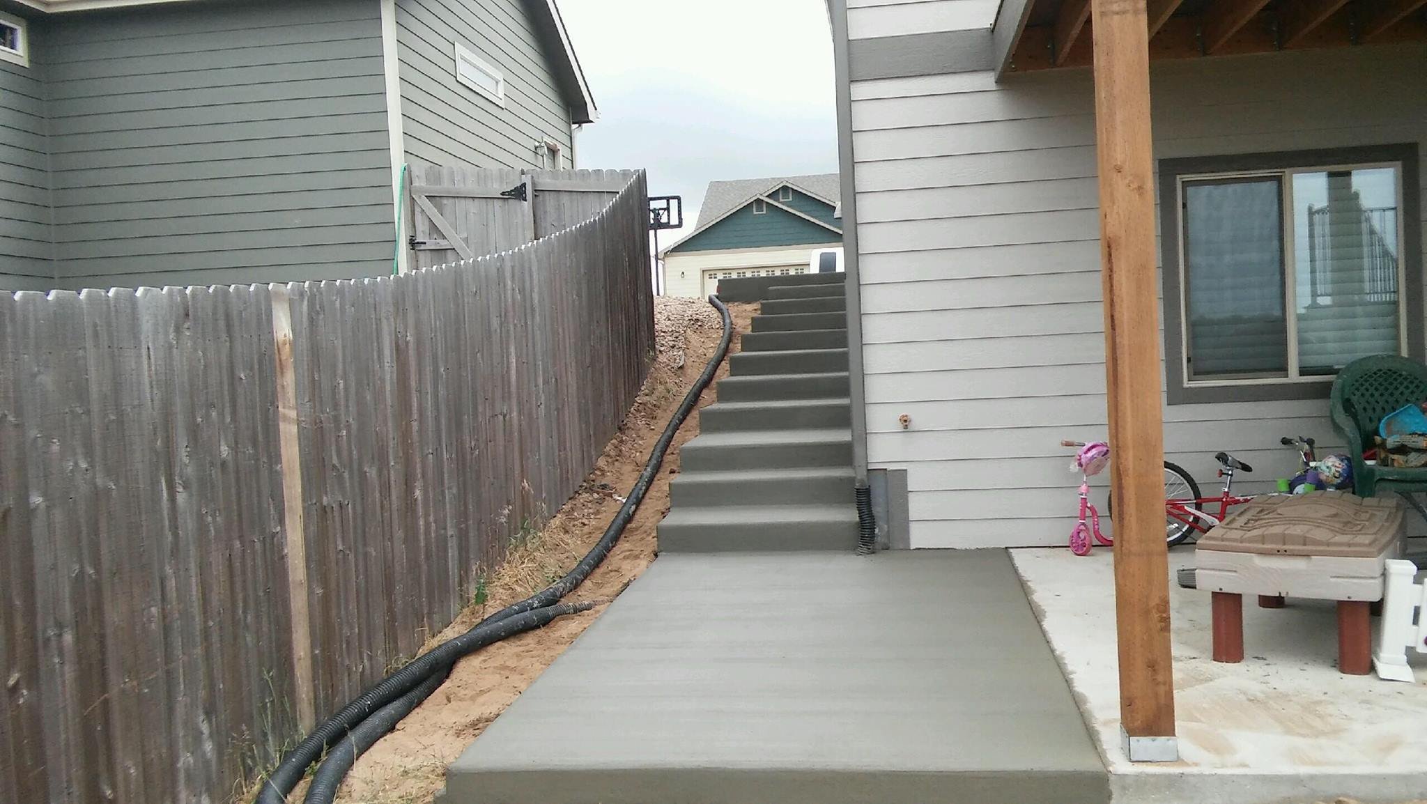 Concrete pathway and stairs beside a house, with a wooden fence on the left and a patio area with children's toys on the right.