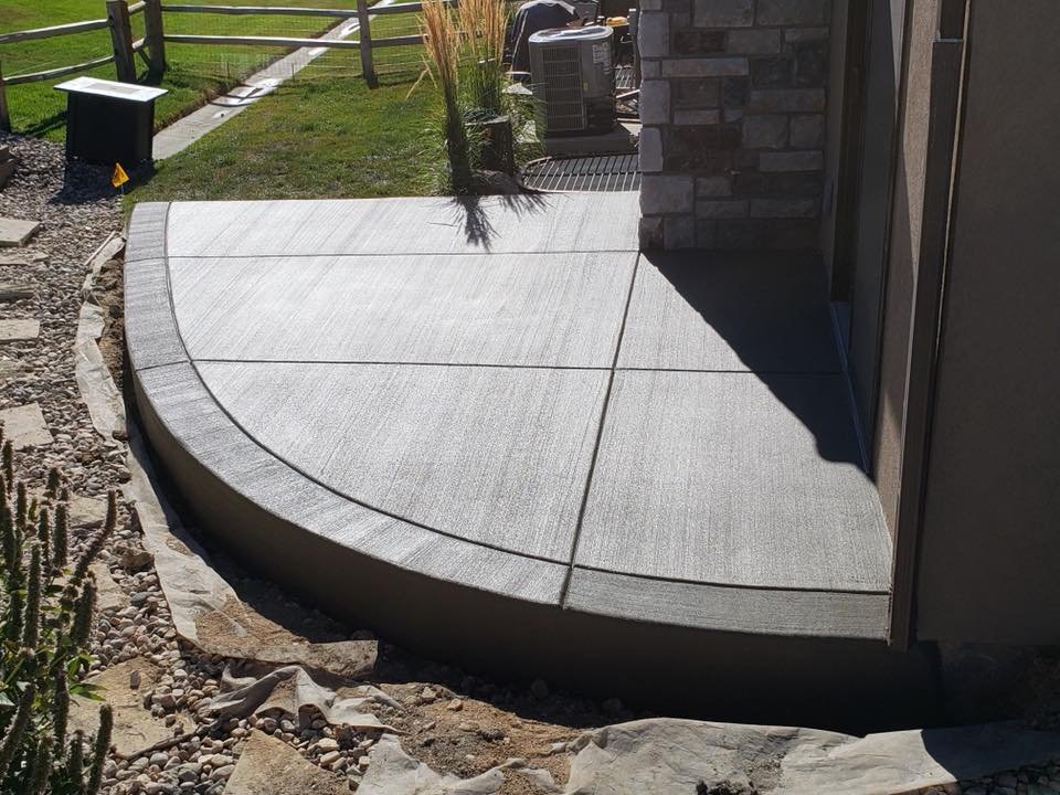 Curved concrete patio with textured finish, adjacent to a building with stone walls and surrounded by gravel and landscaping.