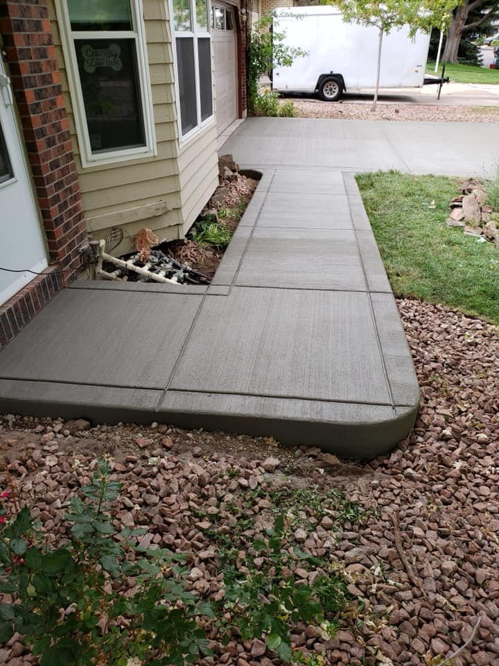 Freshly poured concrete walkway leading to a house with beige siding and a brick exterior. Surrounding landscaping includes red stones and green plants. A white vehicle is visible in the background.