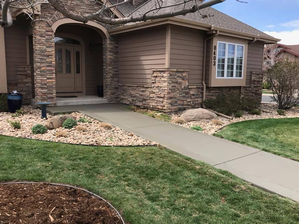 Front exterior of a house with stone facade, manicured lawn, and a concrete pathway leading to the entrance surrounded by landscaped rocks and shrubs.