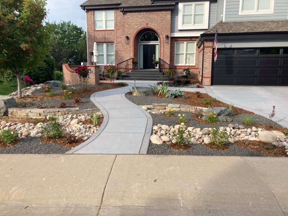 Suburban house with brick facade, landscaped front yard with curved walkway, decorative rocks, plants, and shrubs, two-story structure, American flag near garage.