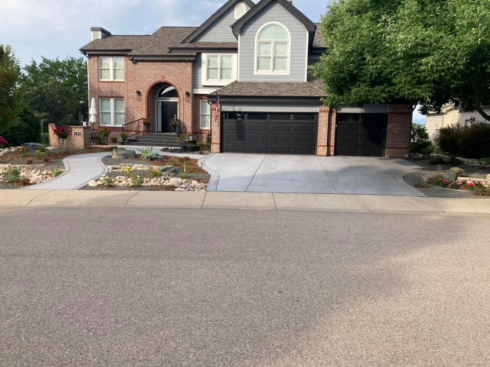 A two-story brick house with a gray roof and black double garage doors, featuring a well-maintained landscaped front yard with a wide concrete driveway and a curved walkway lined with rocks and plants.