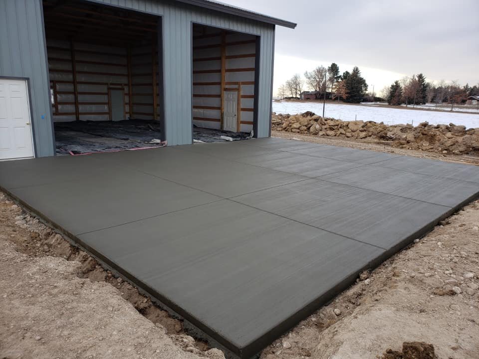 Freshly poured concrete slab in front of a metal building with open bays, surrounded by dirt and construction material.
