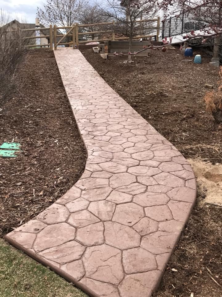 Stamped concrete pathway leading uphill surrounded by mulch with trees and wooden fence in the background.