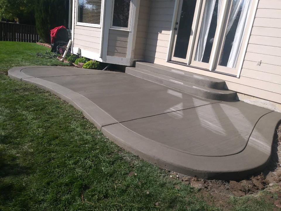 Newly poured concrete patio with curved edges and adjacent grassy lawn.