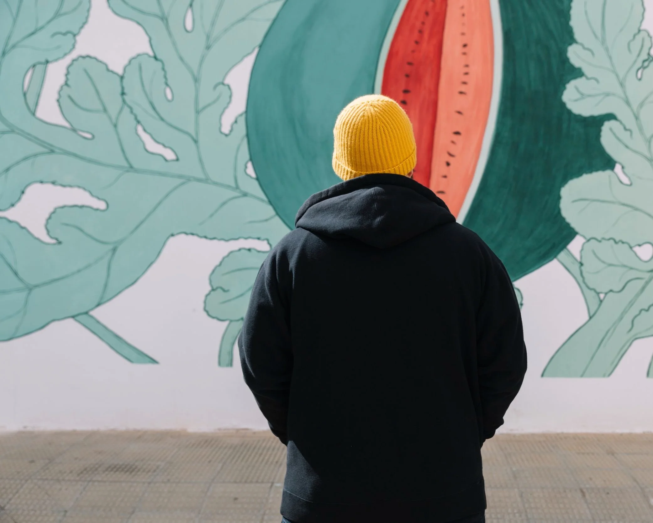 Person in yellow beanie and black hoodie standing in front of a colorful mural with abstract floral design. Sense of Place. Placemaking examples. Unique placemaking