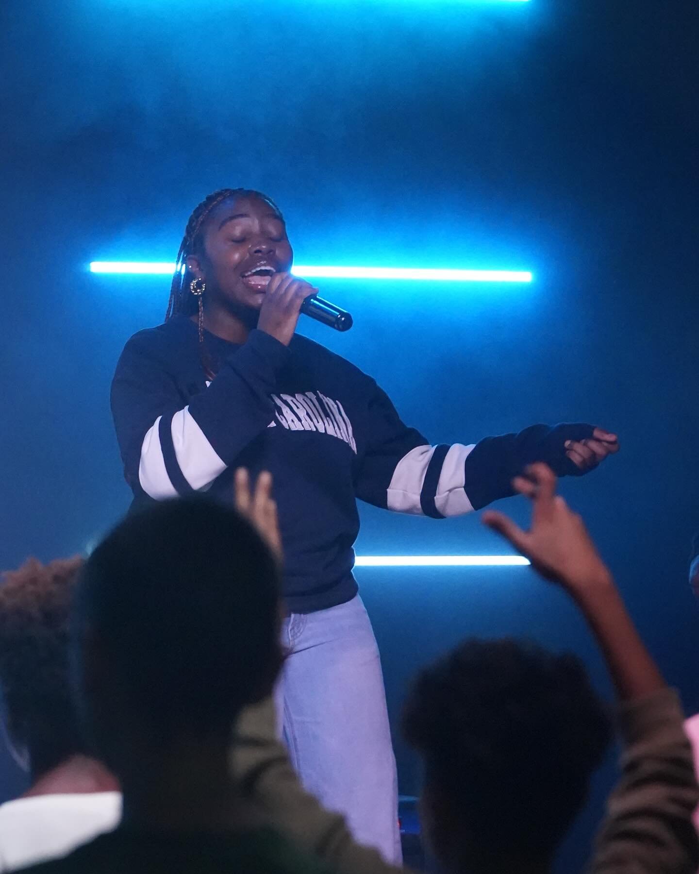 A person singing on stage with a microphone, illuminated by blue lights, surrounded by an audience with raised hands.