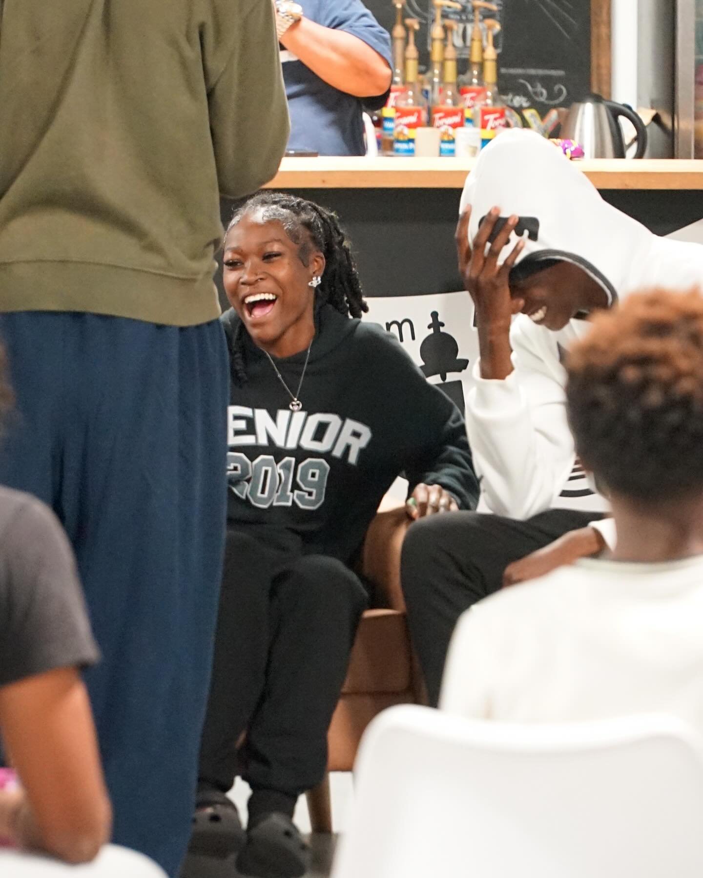 Two people sitting indoors, one wearing a ‘Senior 2019’ sweatshirt, looking happy and engaged. Others are standing or sitting nearby, suggesting a casual group gathering.