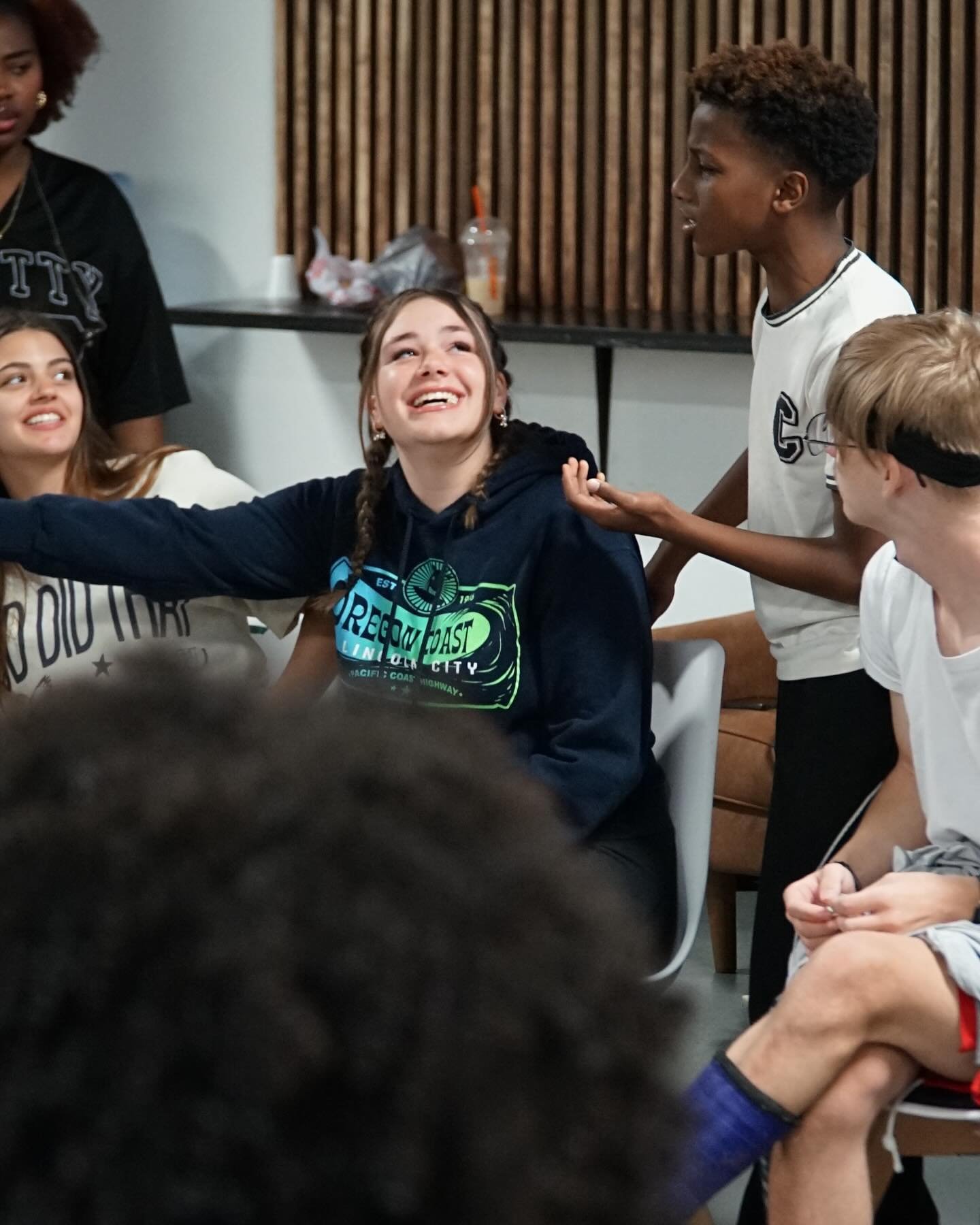 Group of diverse teenagers interacting in a room with wooden paneling and a table with drinks in the background.