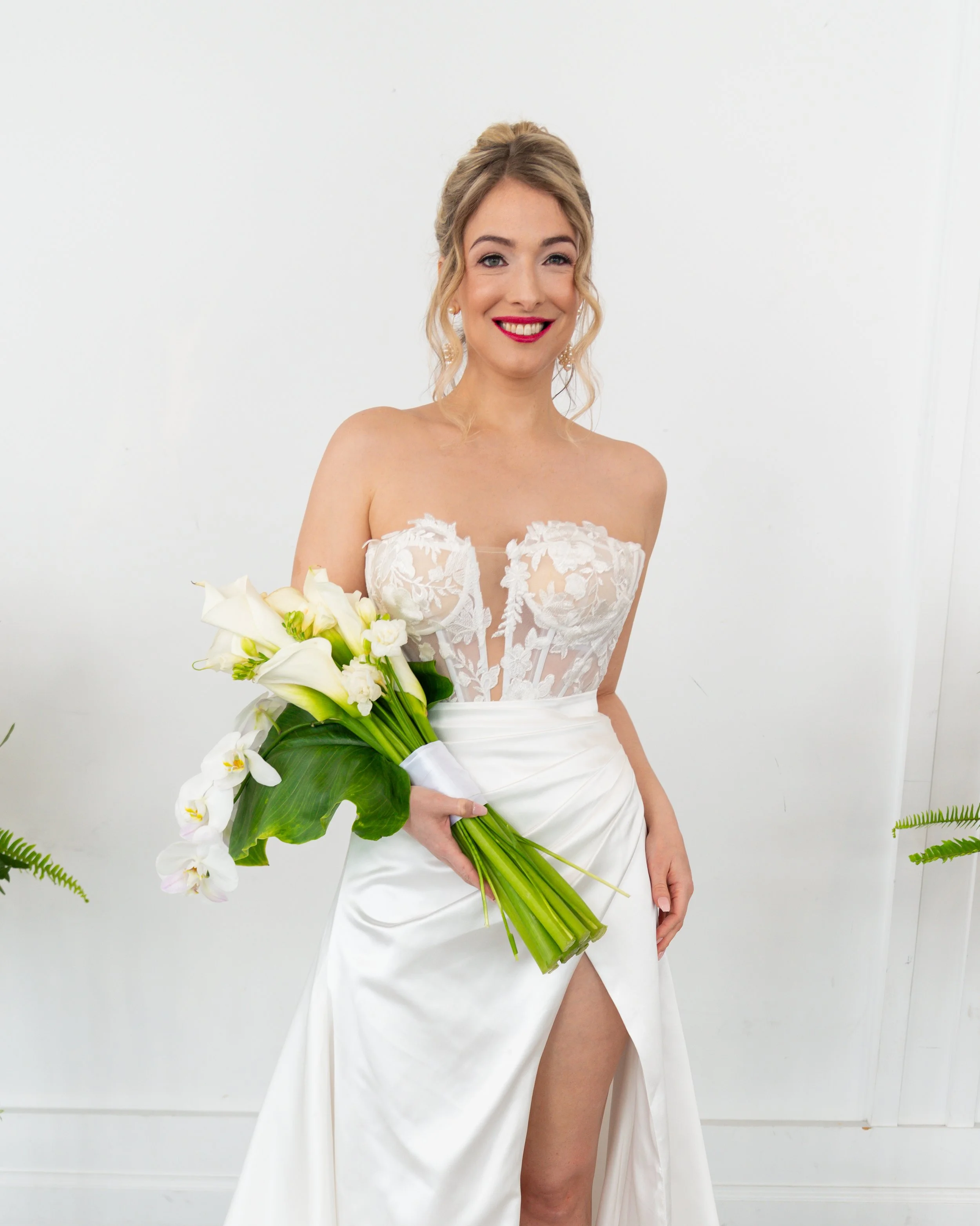 A bride in a white dress holding a large bouquet of white flowers, standing against a plain white wall.