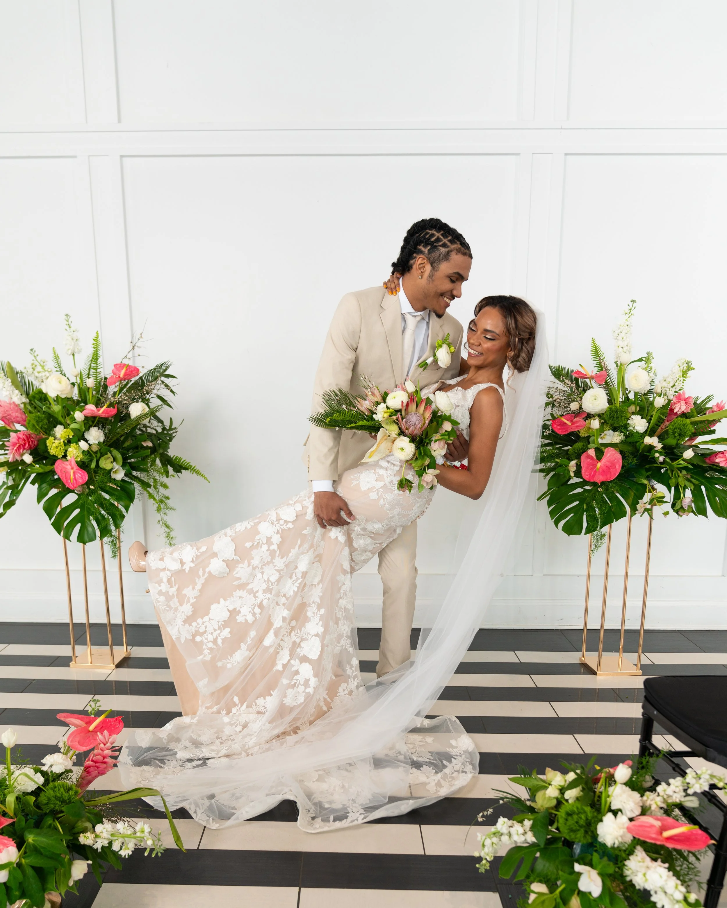 A bride and groom at their wedding, surrounded by floral arrangements, with the groom holding the bride in his arms and smiling at her.