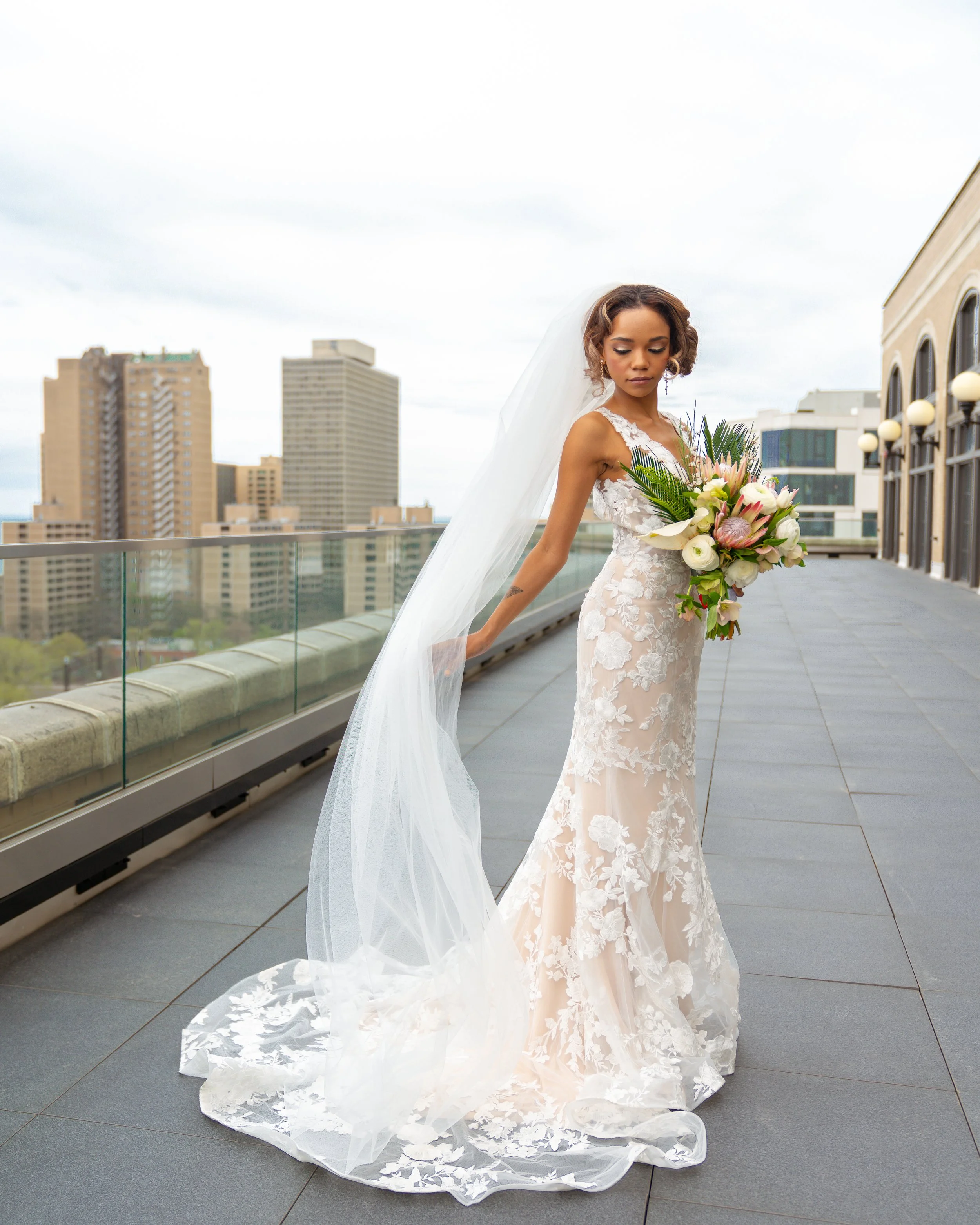 A bride in a white wedding dress with lace details stands on a city rooftop terrace holding a bouquet of white and pink flowers, with buildings and cloudy sky in the background.