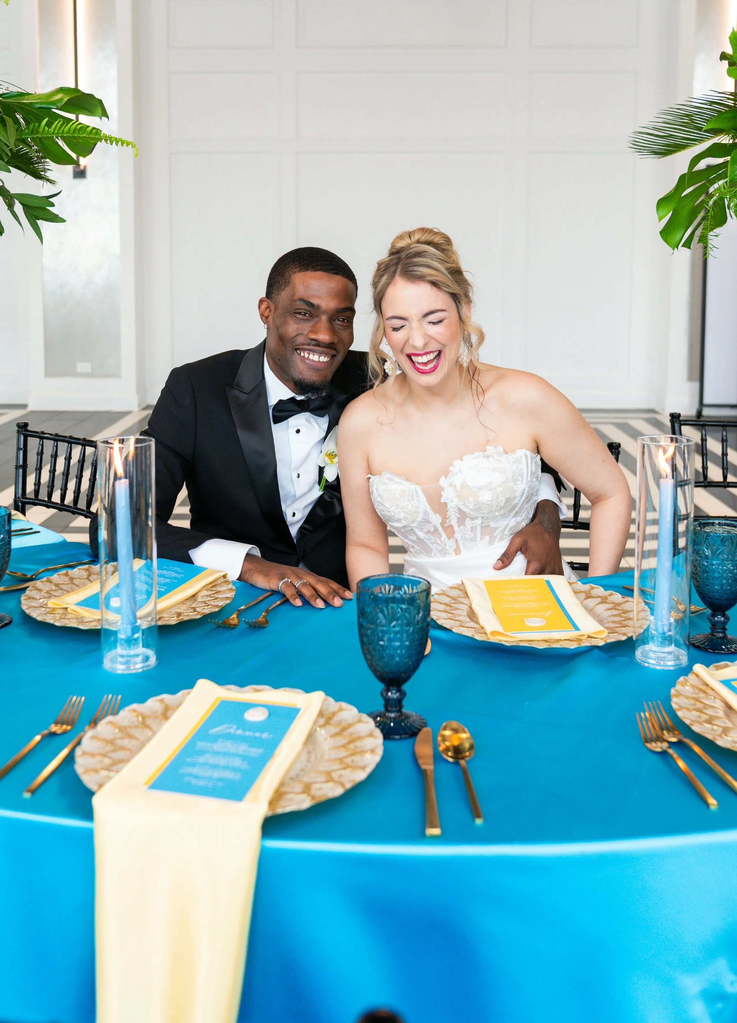 A joyful wedding couple, the bride in a white lace wedding dress and the groom in a black tuxedo, sitting at a vibrant blue dining table with gold accents, surrounded by candles and decorative tableware.