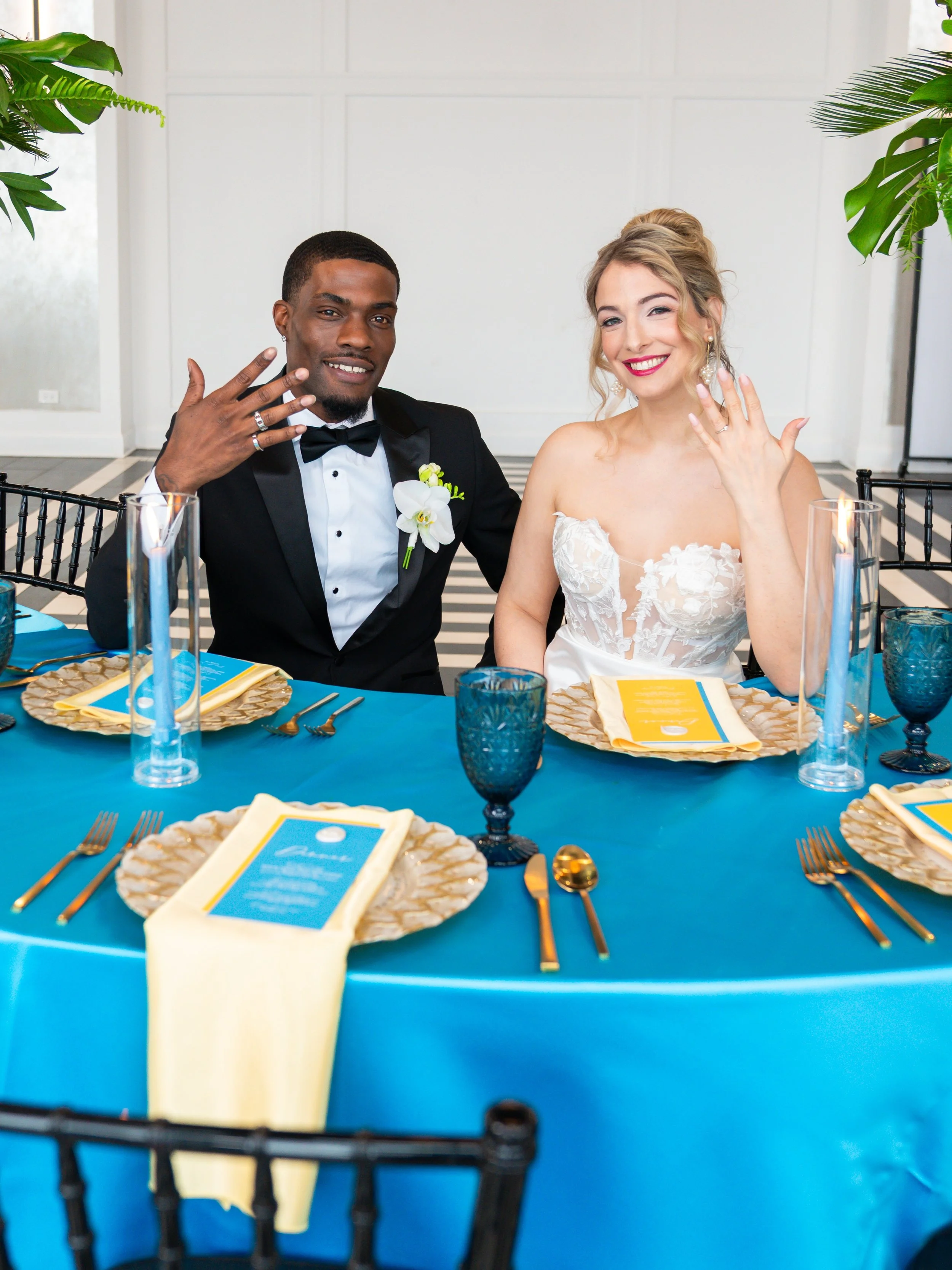 A newlywed couple sitting at a table during their wedding reception, with the groom in a tuxedo and the bride in a wedding dress, both showing rings and smiling.