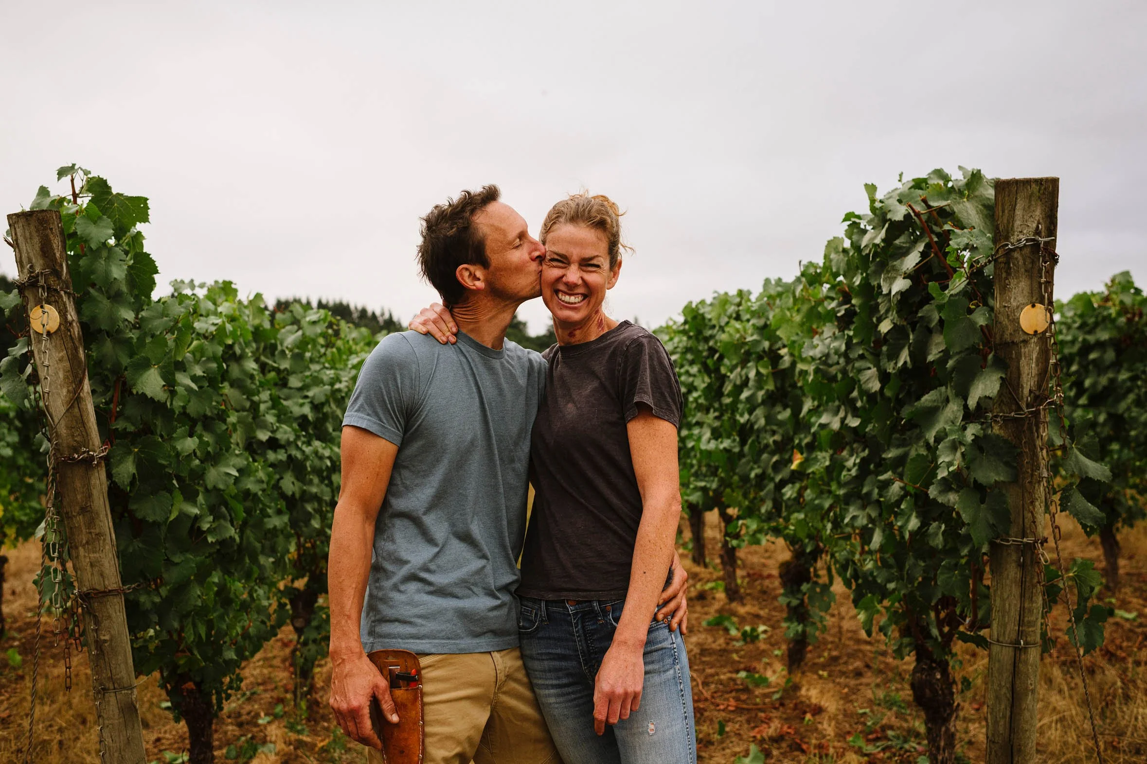 A man and woman stand close together in a vineyard, smiling and showing affection, with grapevines in the background.