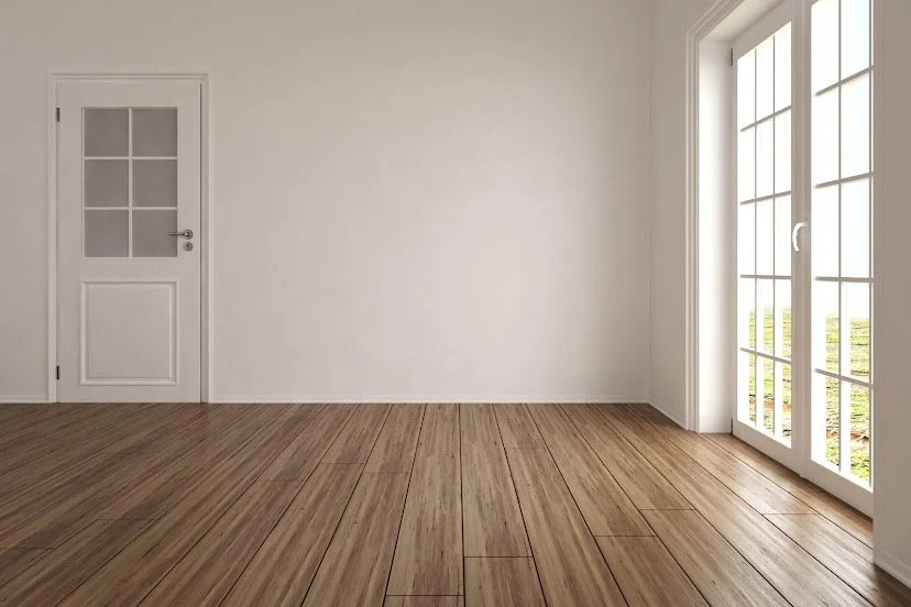 Empty room with white walls, wooden flooring, a white door with glass panels, and large windows with white frames allowing natural light to enter.