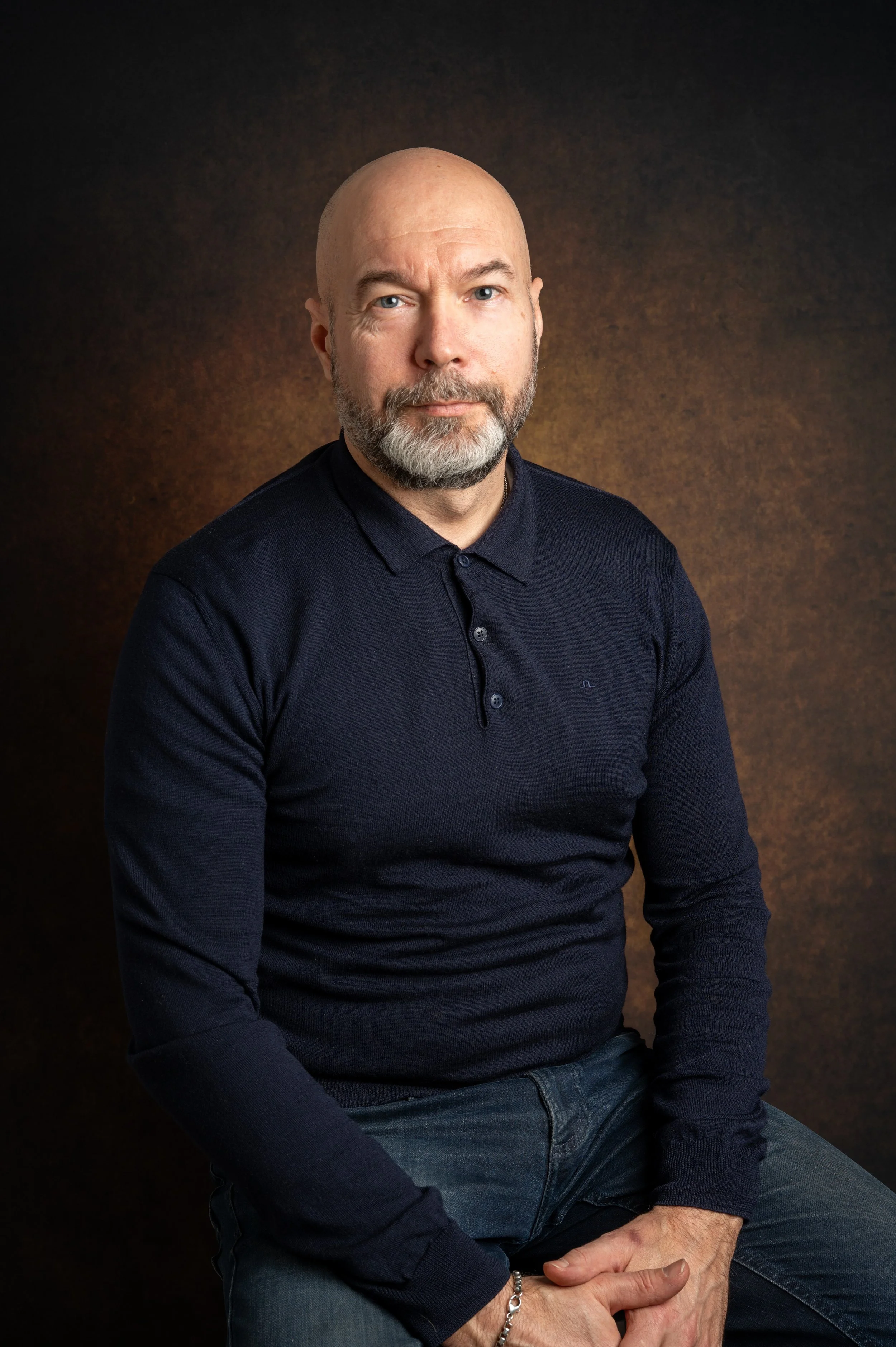 Mies istuu mustavalkoisessa valokuvassa, pukeutuneena tummaan pikeepuseroon ja farkkuihin, taustalla ruskea sävy.