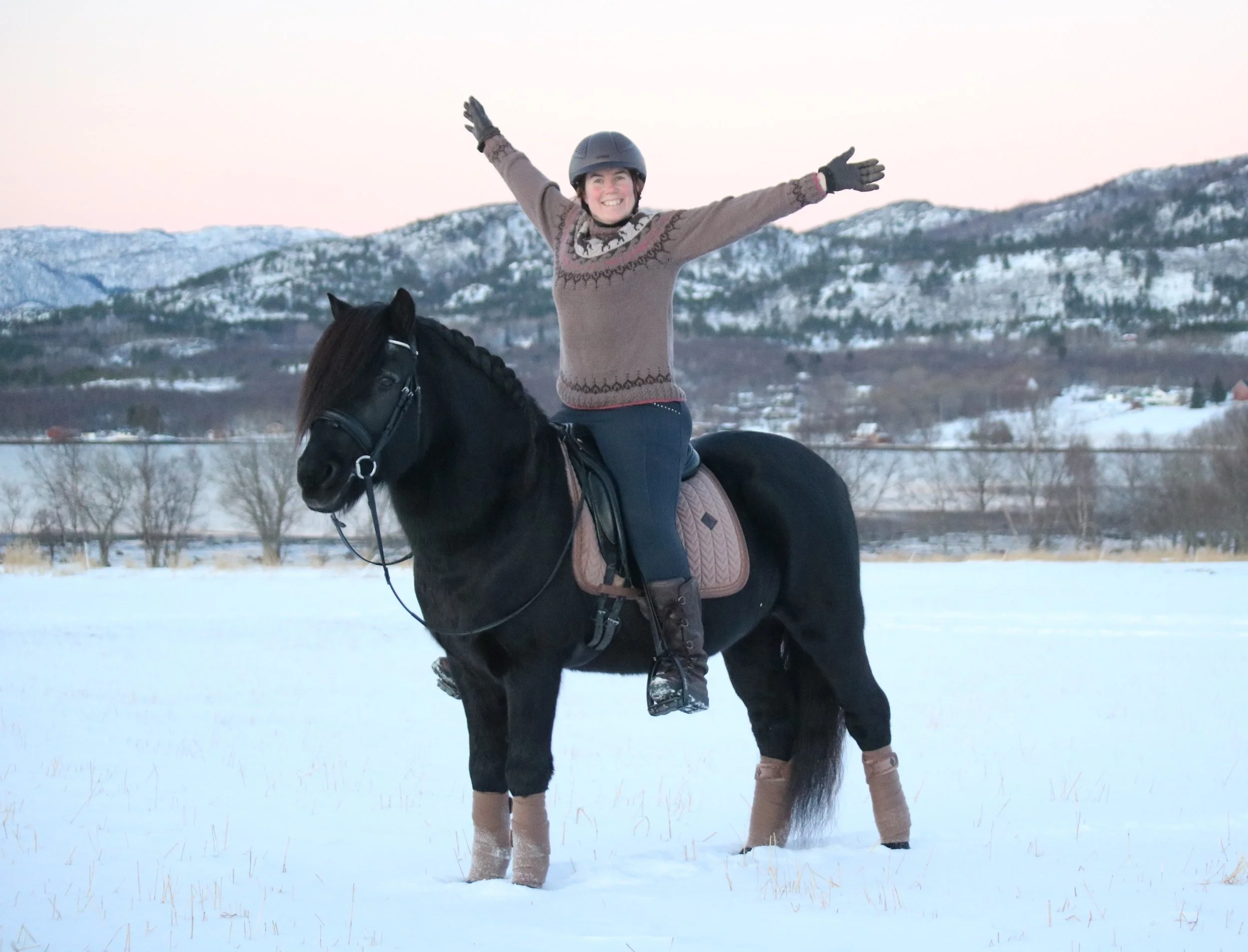En kvinne rir på en svart hest i snølandskap med fjell i bakgrunnen, hun smiler og har armene utstrakt.